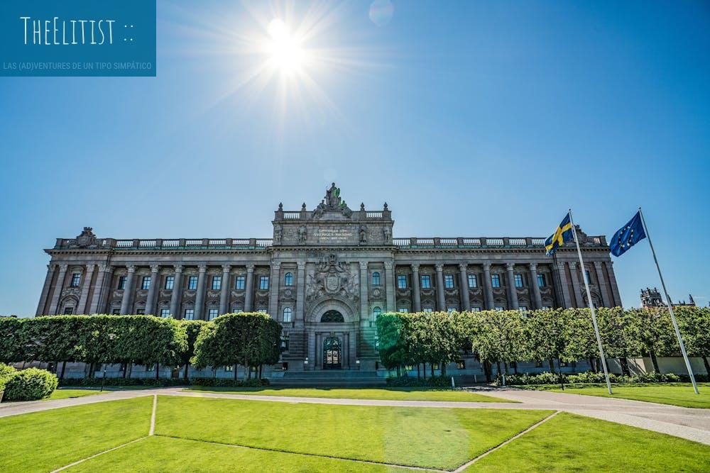 Front view of the Riksdag building with Swedish and EU flags on a sunny day.
