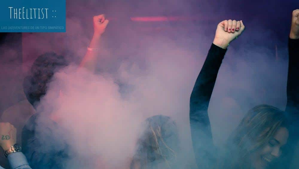 Crowd of people dancing energetically in a foggy nightclub with raised hands.